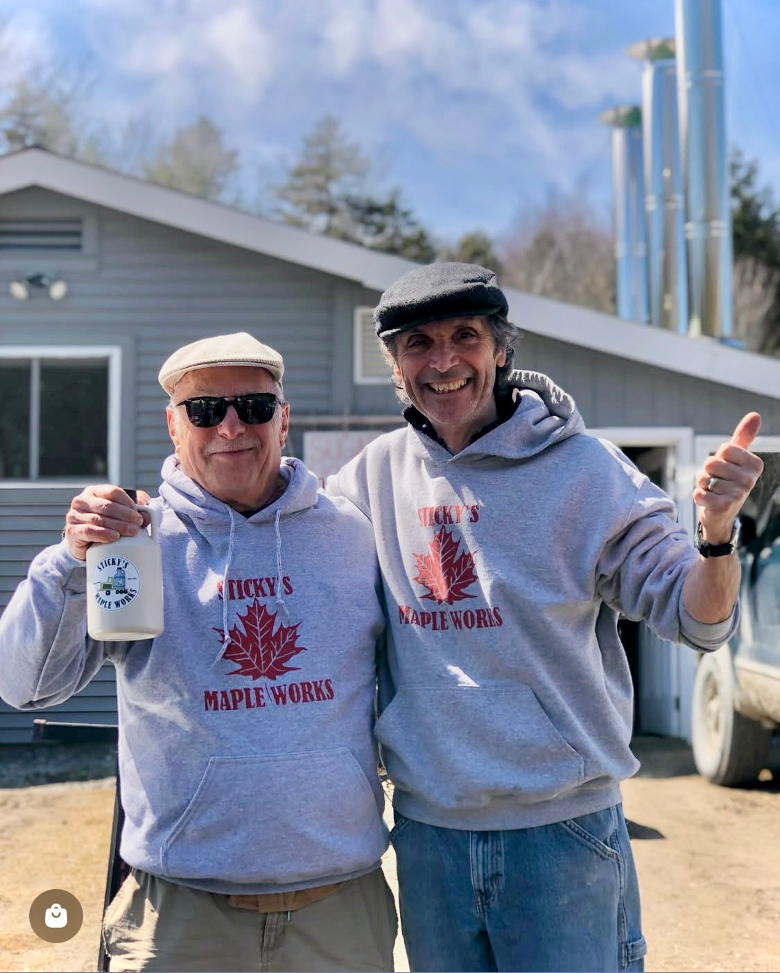 Two men wearing 'Sticky's Maple Works' hoodies, one holding a mug, outdoors with a building and trees in the background.